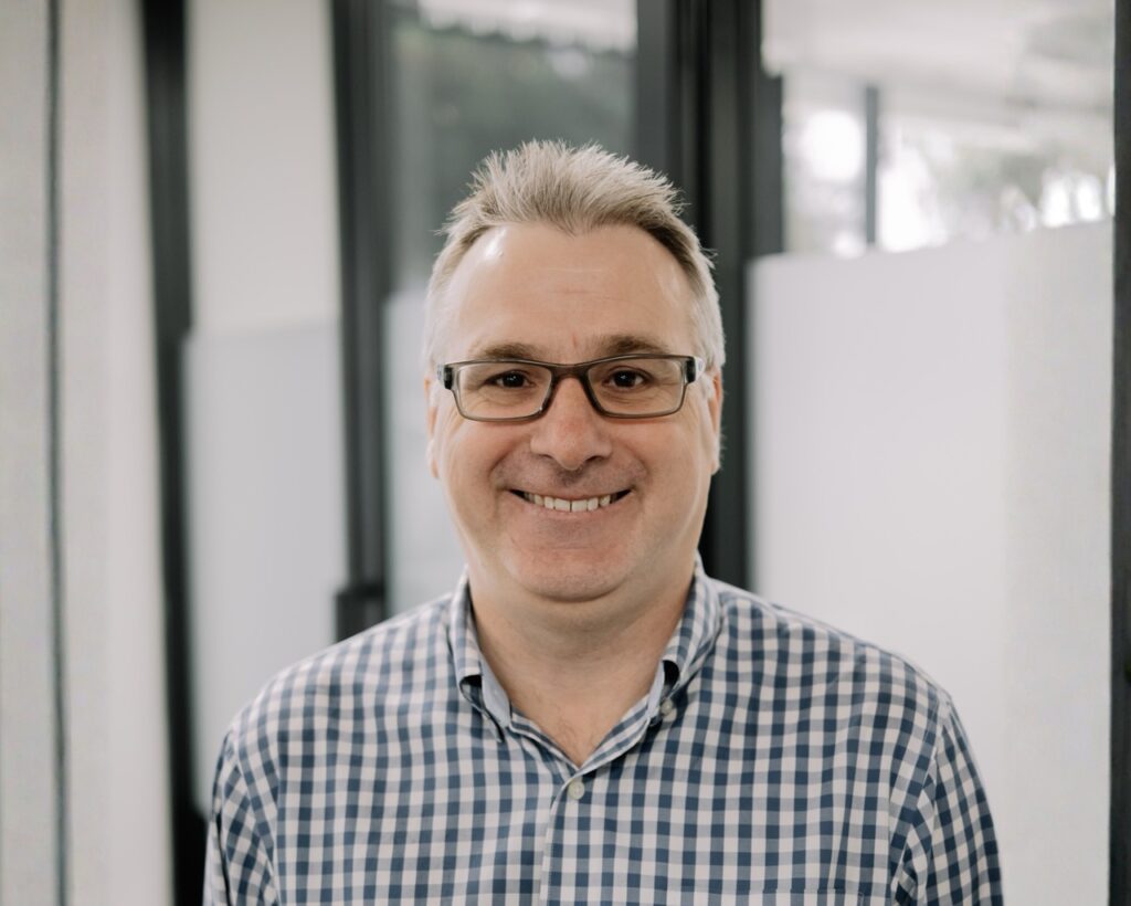 A man with short grey hair and glasses, wearing a blue and white checked shirt, smiling whilst standing indoors in front of glass panels.