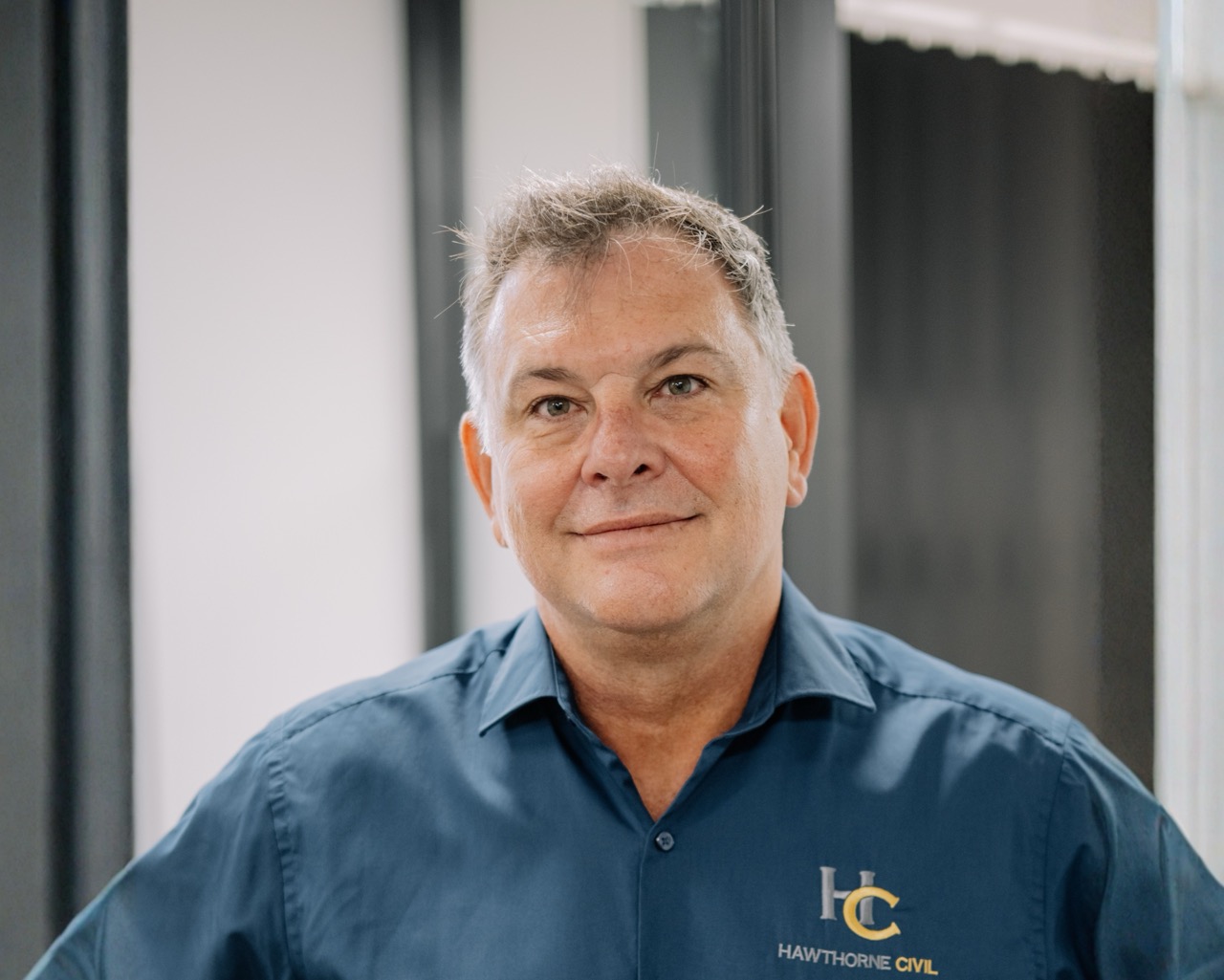 A middle aged man with short grey hair, wearing a blue shirt with a Hawthorne Civil logo, stands indoors in front of a blurred office background, smiling slightly at the camera.