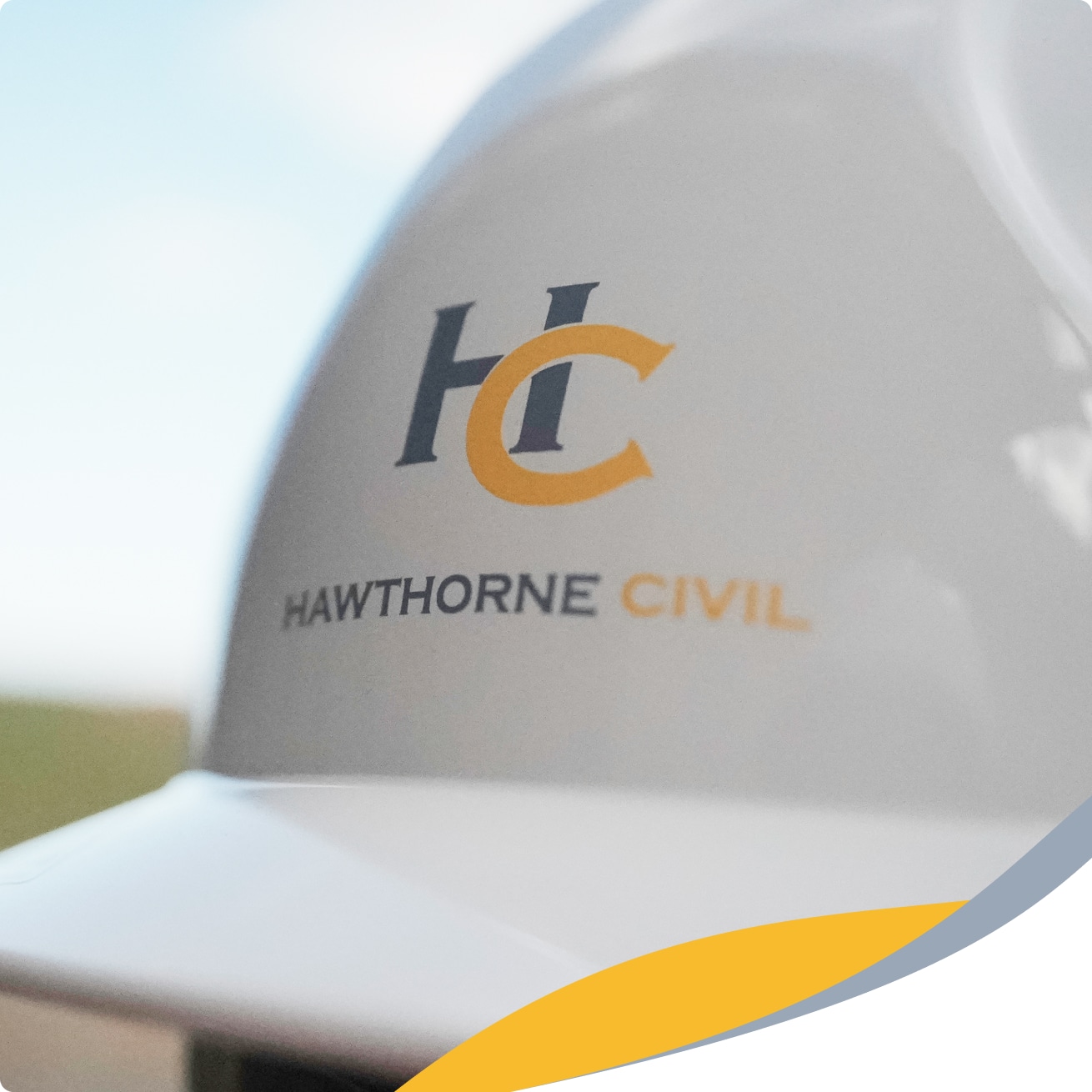 A close-up of a white construction hard hat featuring the HC logo and the text HAWTHORNE CIVIL in grey and gold letters, with a blurred outdoor background.