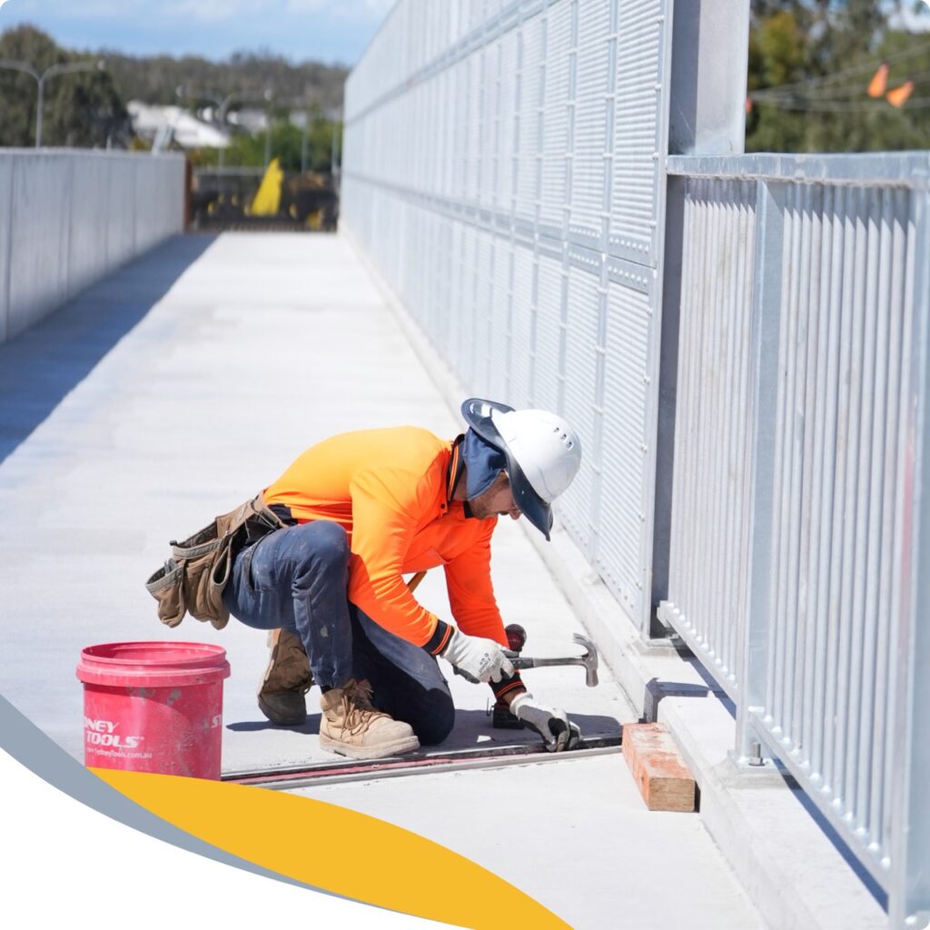 A construction worker in a bright orange shirt and hard hat kneels on a concrete bridge, using a hammer and tool to work on a metal railing. A red bucket and bricks are nearby, with safety equipment visible.