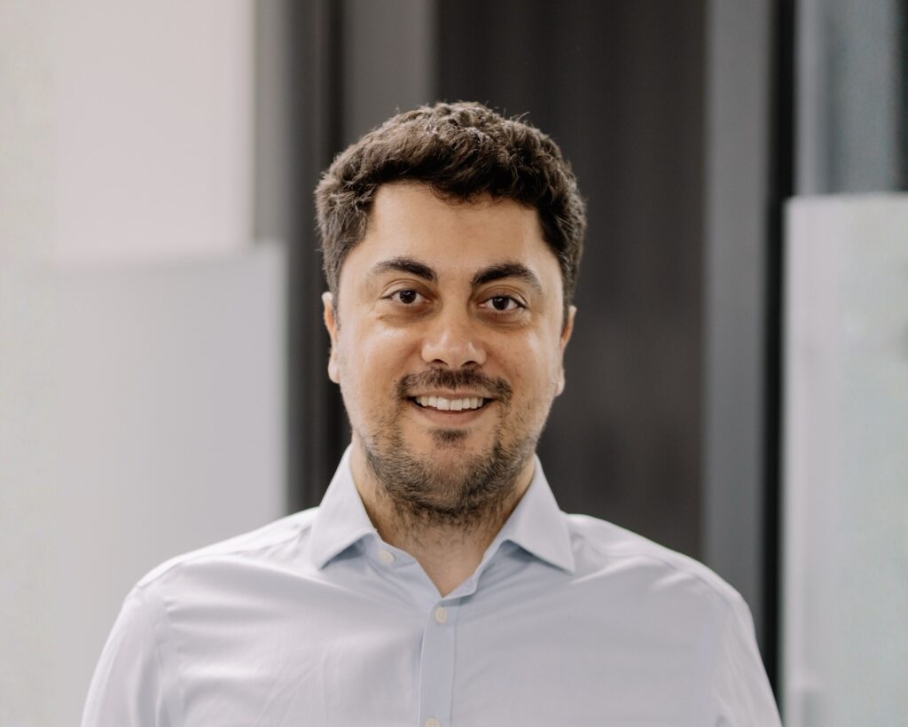 A man with short dark hair and facial hair is smiling at the camera. He is wearing a light blue button up shirt and is standing indoors against a blurred background.