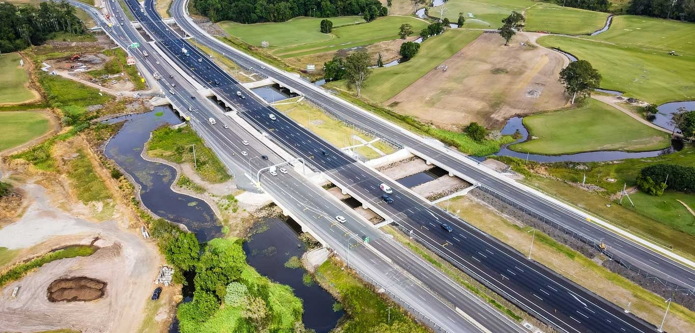 Aerial view of a large multi lane motorway interchange surrounded by green fields, trees, and a few ponds, with cars travelling in both directions.