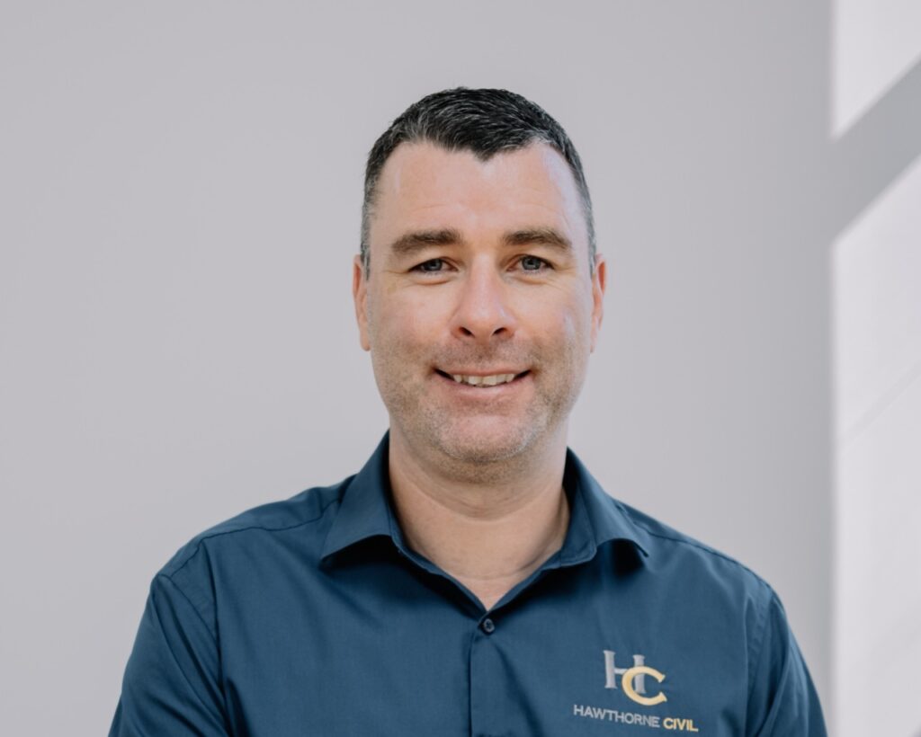 A man with short dark hair smiles at the camera. He is wearing a dark blue collared shirt with the logo and text HC Hawthorne Civil on it. The background is plain and light coloured.
