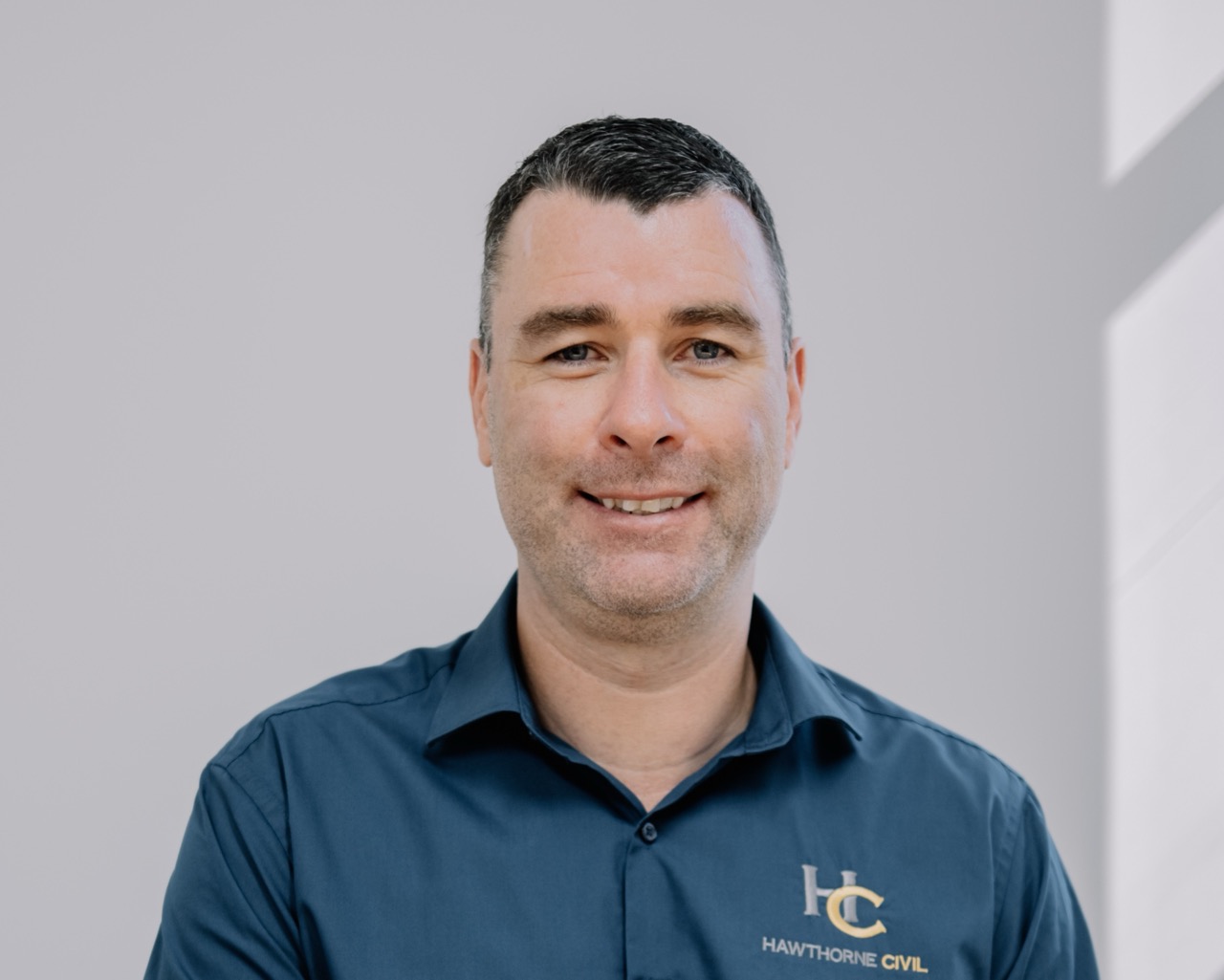A man with short dark hair smiles at the camera. He is wearing a dark blue collared shirt with the logo and text HC Hawthorne Civil on it. The background is plain and light coloured.