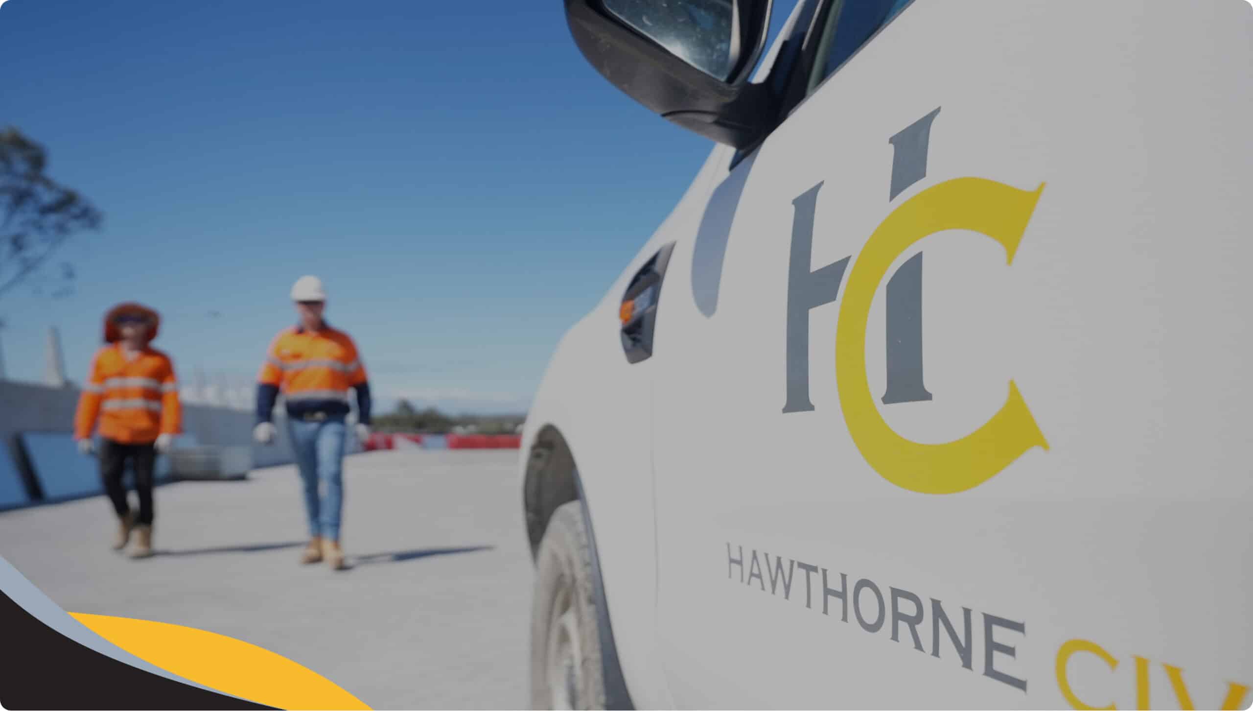 Two construction workers in orange vests and helmets walk on a building site; the foreground shows a white vehicle with the Hawthorne Civil logo and the letters HC on the side.