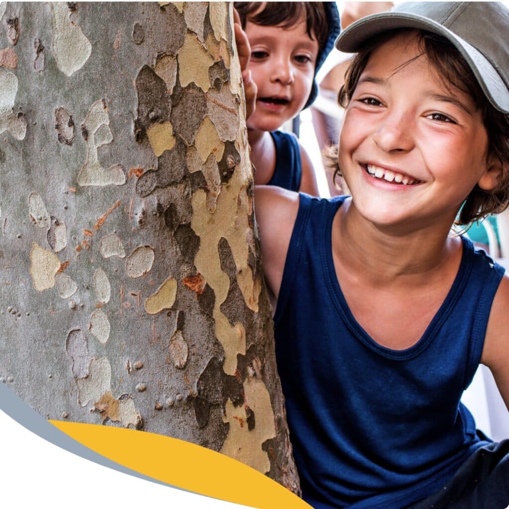 Two children in blue vests smile and peek out from behind a tree with textured bark. One wears a grey cap, and both seem happy and playful outdoors.