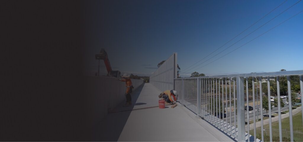 Two construction workers are working on a rooftop walkway near a metal railing under a clear blue sky, with one kneeling and the other standing. The right side shows a view of the surrounding area.