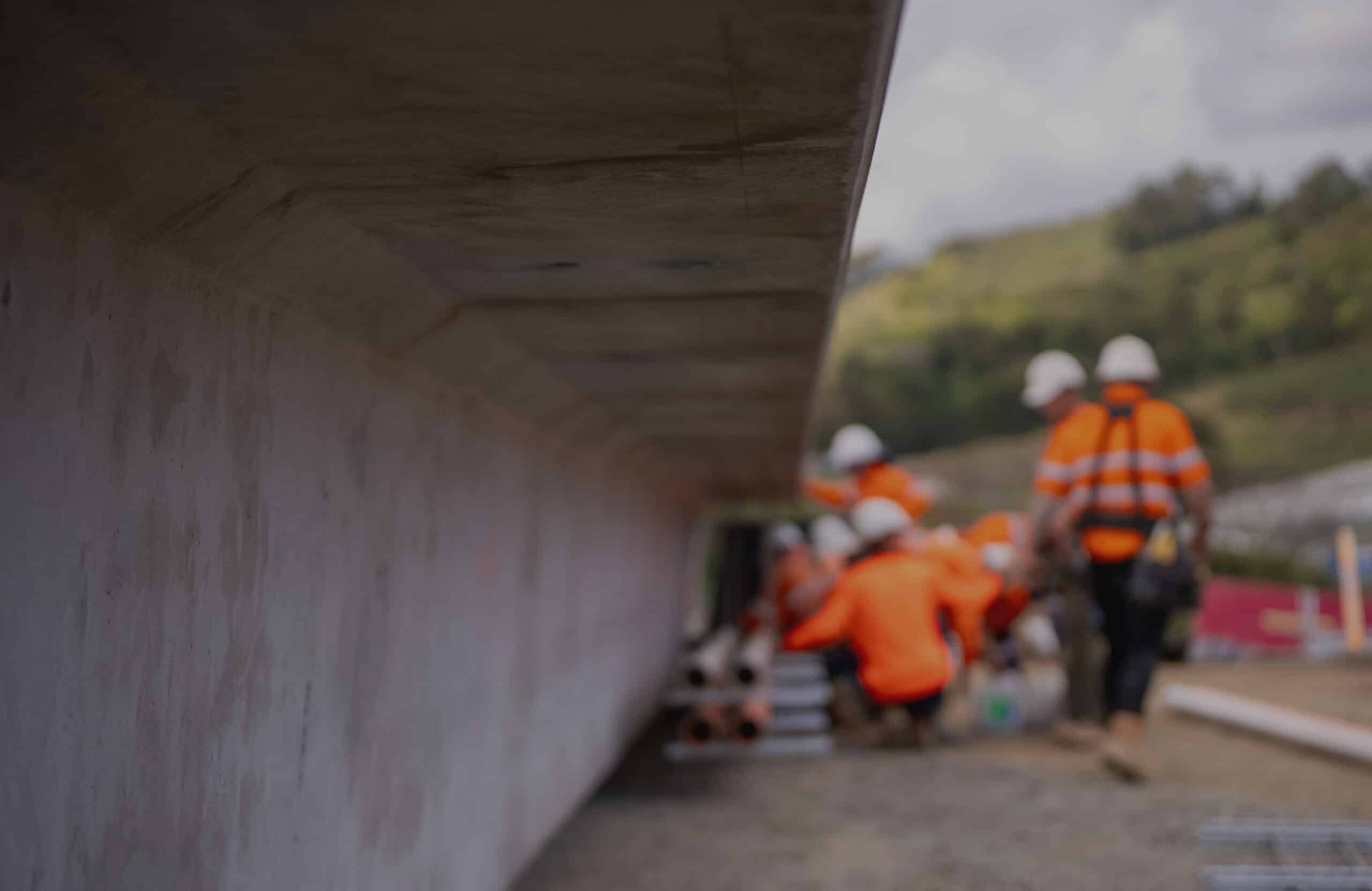 A group of construction workers in orange safety vests and helmets work together near a large concrete slab outdoors, with trees and hills in the background. The image is focused on the slab with the workers blurred.