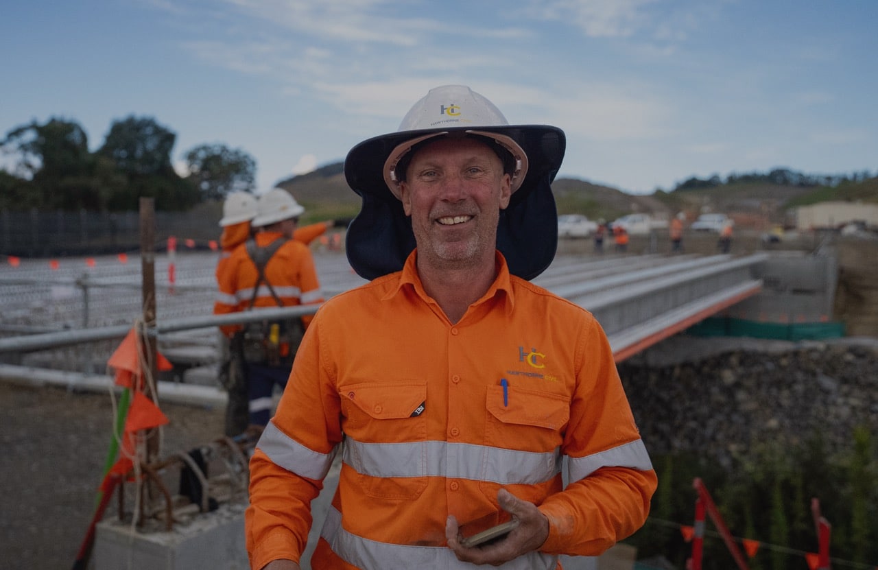 A construction worker in an orange high-visibility shirt and white hard hat smiles at the camera at a bridge construction site. Other workers and construction materials are visible in the background.