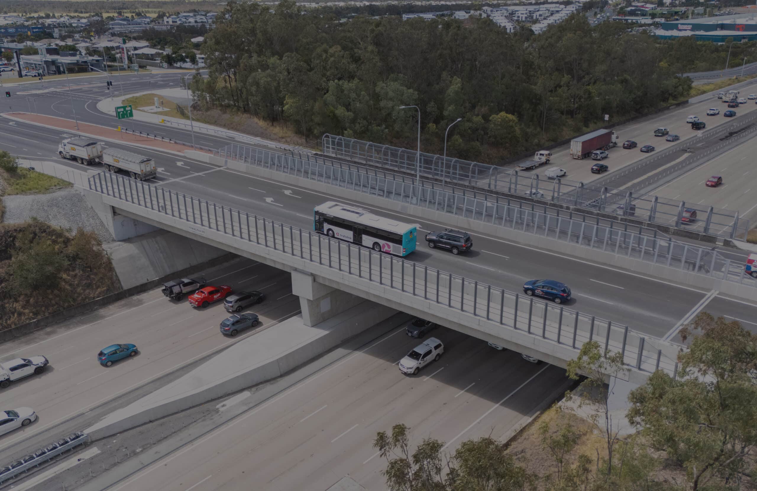 An aerial view of a motorway flyover with cars, a lorry, and a bus travelling on the upper road, and several vehicles moving on the road below, surrounded by trees and urban areas.