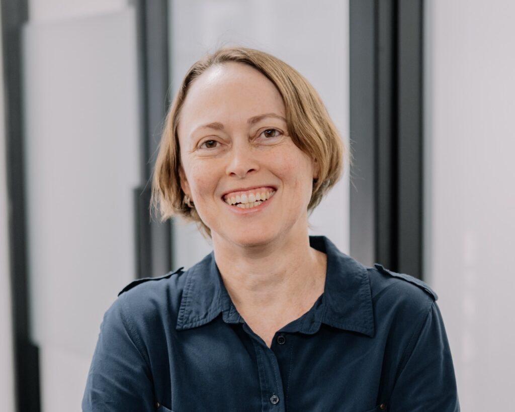 A woman with short light brown hair, wearing a dark blue collared shirt, smiles warmly at the camera. She is indoors, with blurred glass panels in the background.