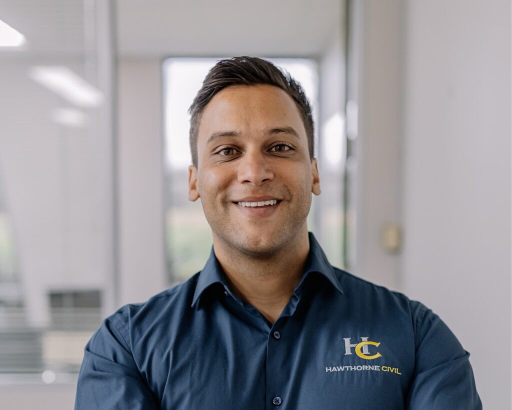 A smiling man with short dark hair wearing a navy blue Hawthorne Civil shirt stands in a modern office space with blurred windows and glass walls in the background.