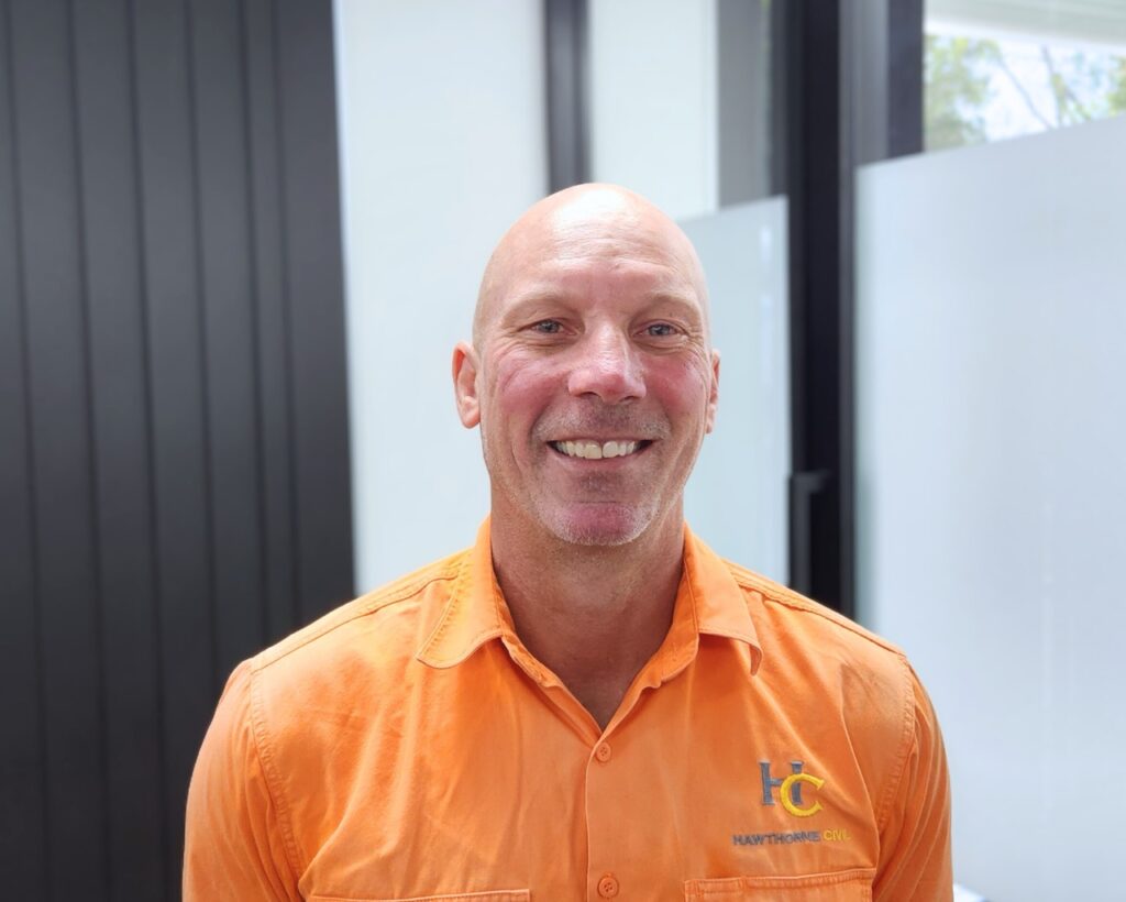 A smiling man in an orange collared shirt with an HC logo stands indoors in front of frosted glass panels and a dark wall.