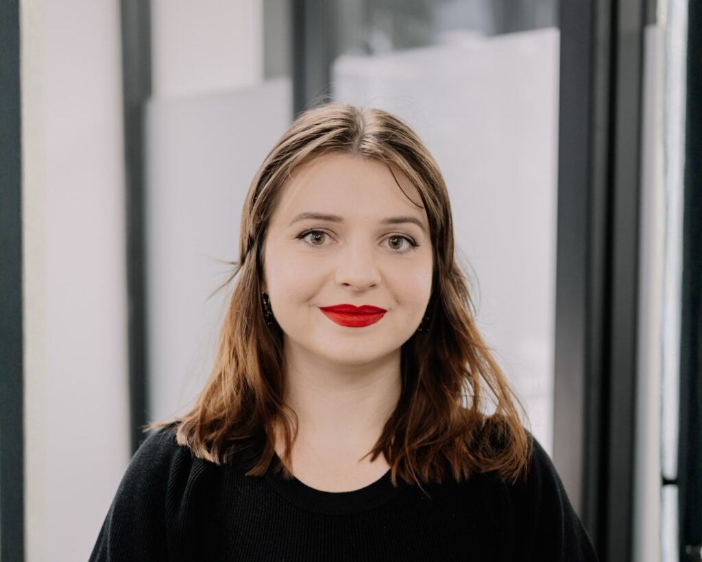 A young woman with shoulder length brown hair and bright red lipstick is standing indoors, facing the camera and smiling softly. She is wearing a black top, and the background features large windows and blurred office elements.