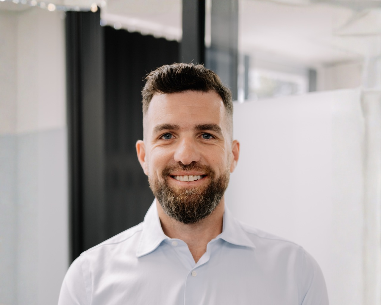 A man with short brown hair and a beard, wearing a light blue collared shirt, smiles at the camera in a bright, modern indoor setting with blurred windows and white walls in the background.
