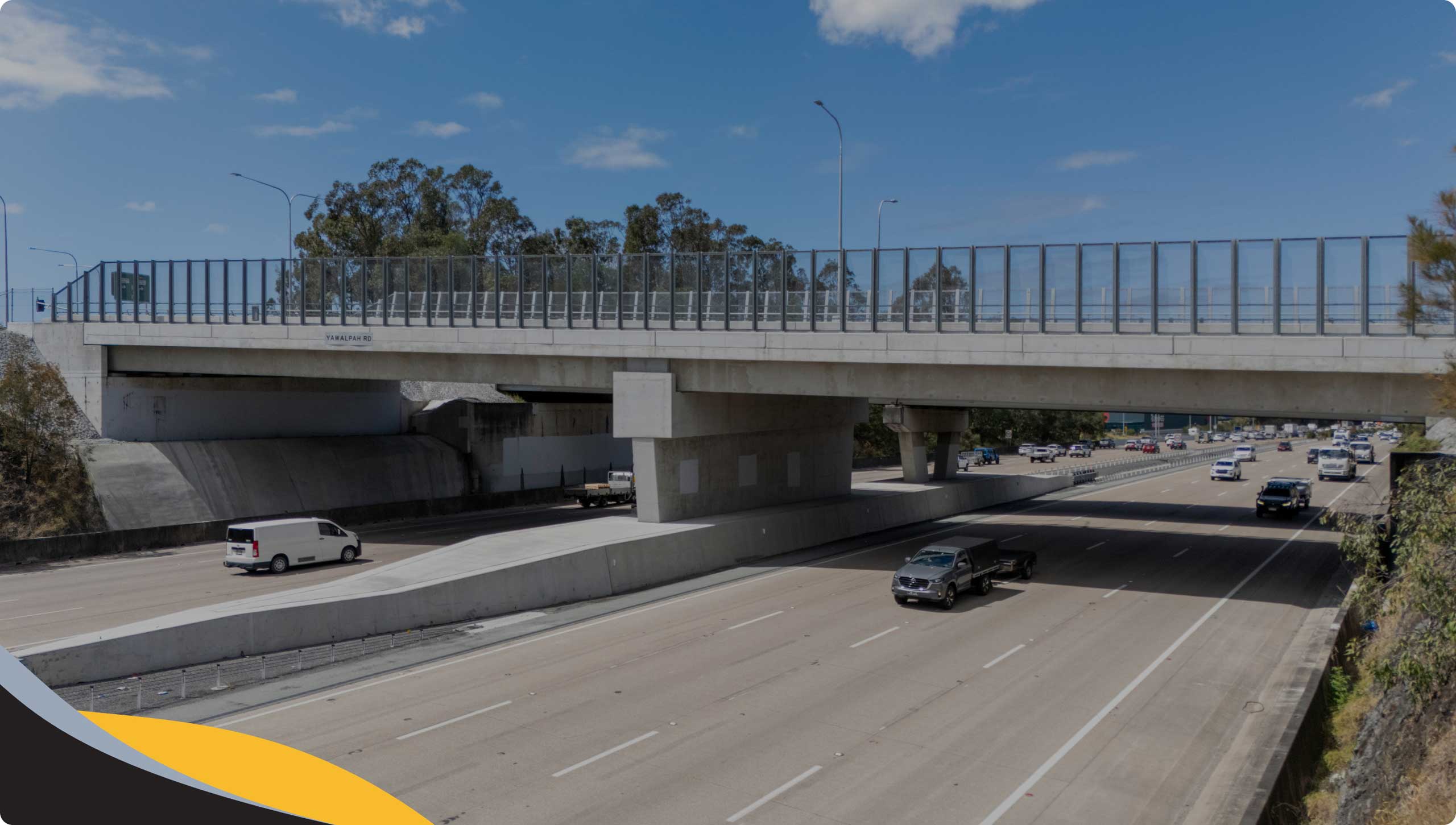 A concrete flyover with glass barriers spans across a multi-lane motorway with vehicles travelling in both directions under a blue sky with scattered clouds. Trees and foliage line the sides of the motorway.
