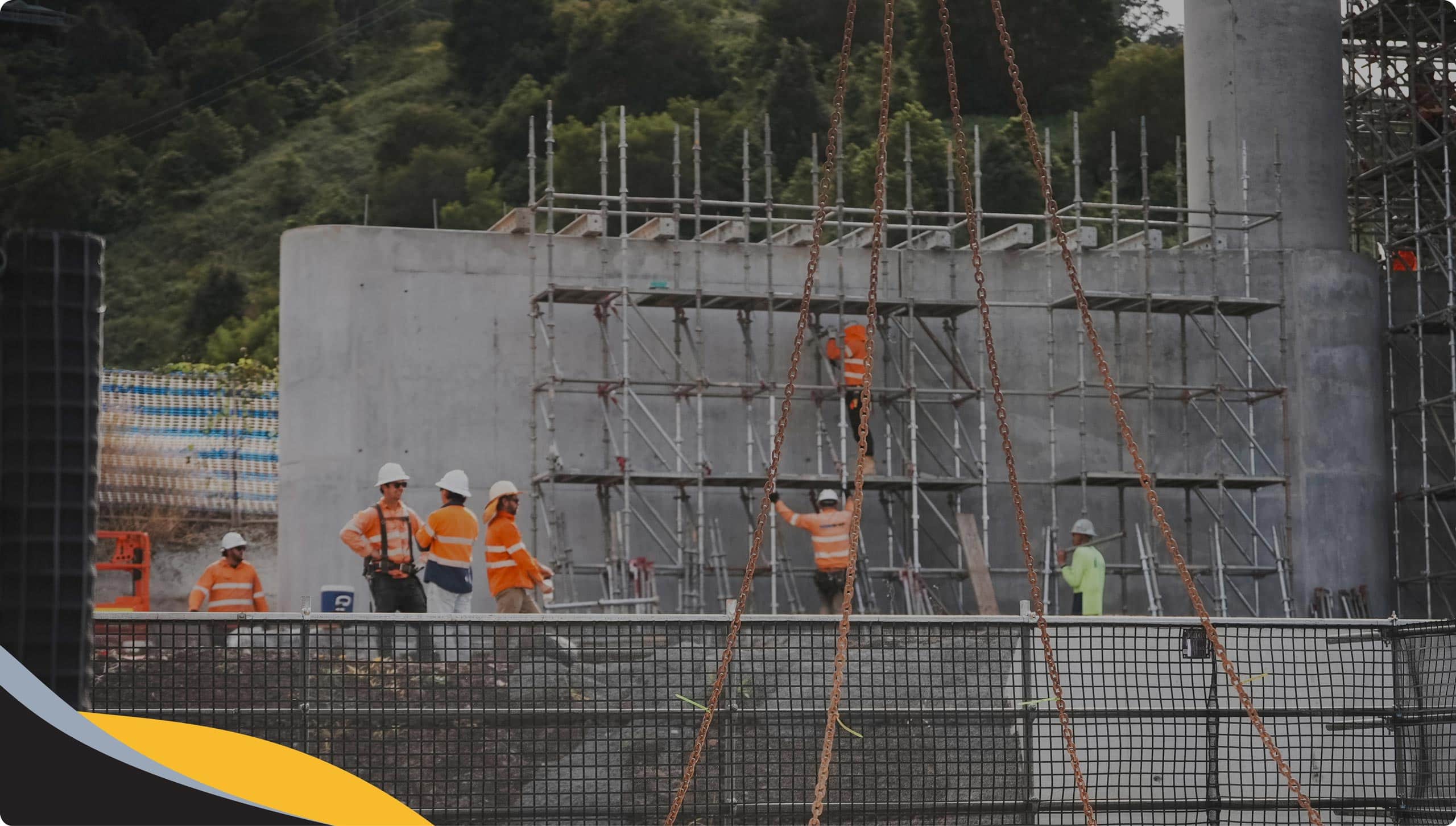Construction workers wearing safety gear stand and work on scaffolding and a concrete structure at an outdoor construction site, surrounded by greenery and building materials.