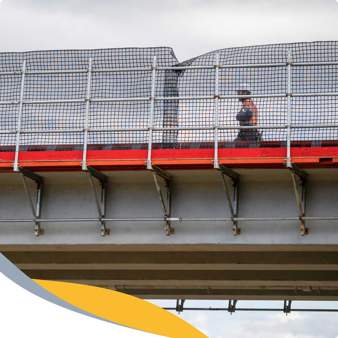 A person wearing a hard hat and safety vest walks along a fenced walkway on a bridge. The fence is slightly bent near the top, and the bridge structure is visible below.