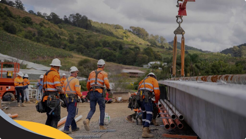 Construction workers in orange safety vests and helmets work together near a large structure with metal pipes, surrounded by greenery and hills under a cloudy sky. A crane hook is visible above the site.