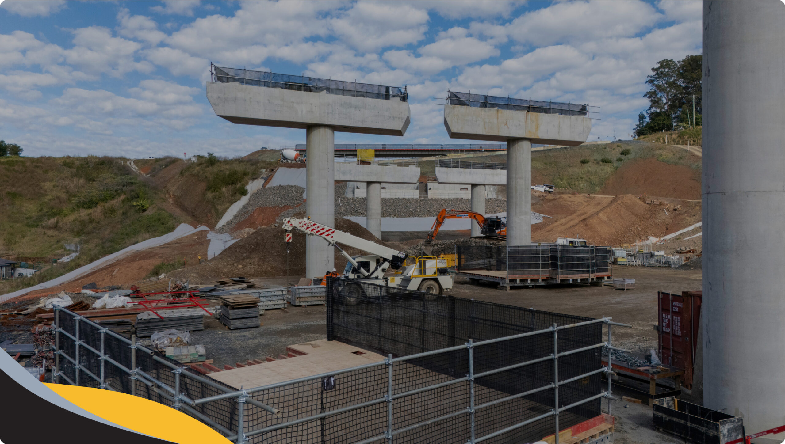 A construction site with large concrete bridge supports, cranes, and building materials. The area is surrounded by soil, hills, and fencing under a partly cloudy sky.