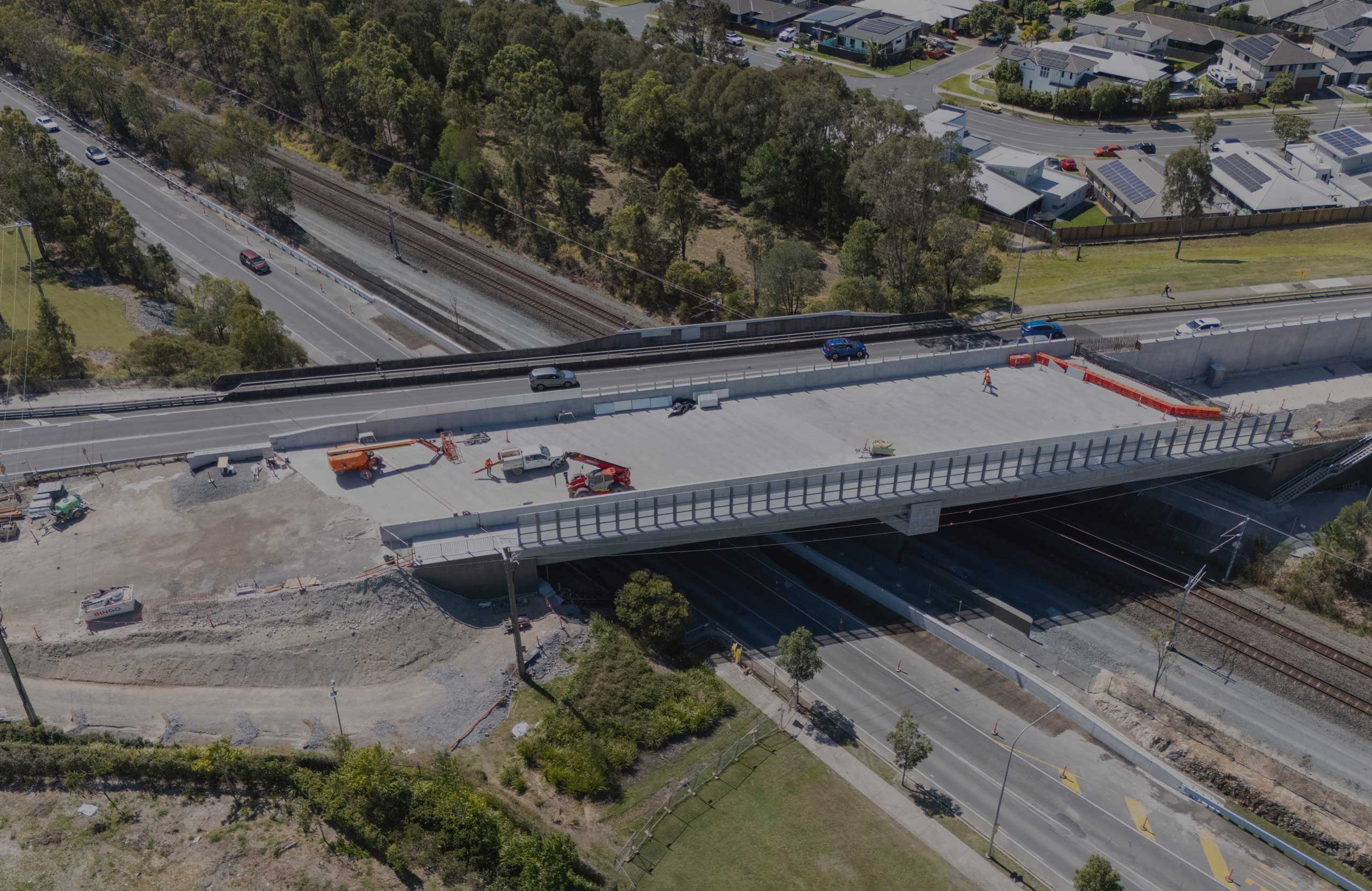 Aerial view of a large concrete bridge under construction over a road and railway, with construction vehicles and workers present, surrounded by trees and residential houses.