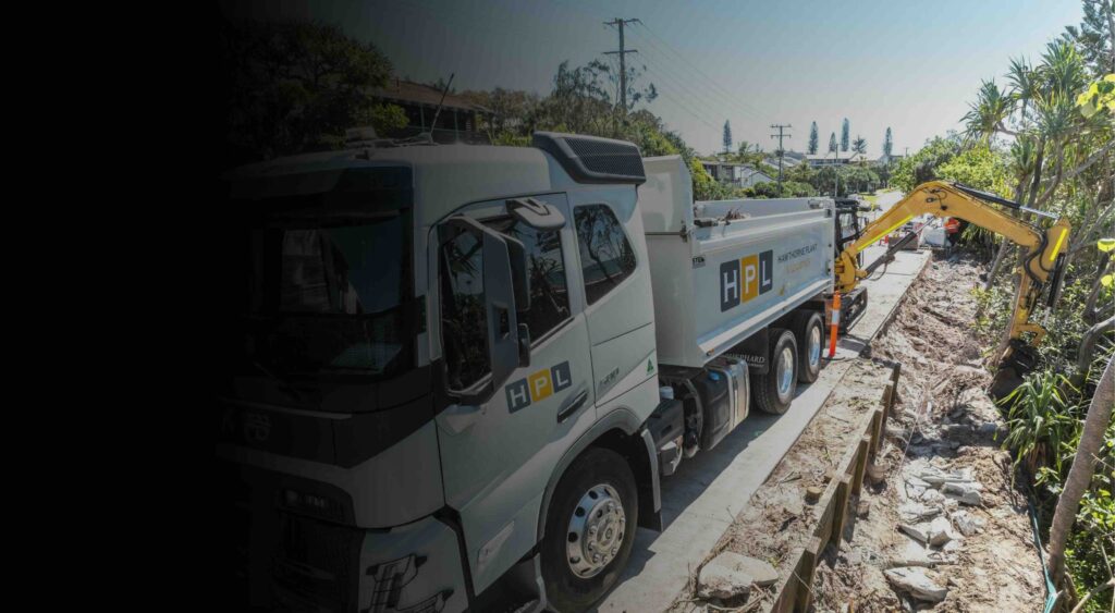 A white HPL branded construction lorry is parked on a street beside an excavator digging a trench, with trees and utility poles lining the road.